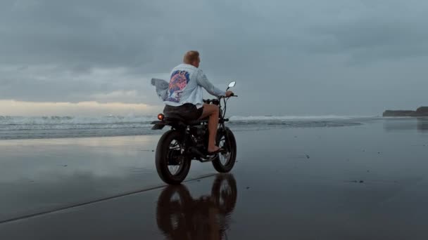 Ung stilig hipster man rider modern anpassad motorcykel racer på den svarta sandstranden nära vattnet. Surfingplats med havsvågor. — Stockvideo