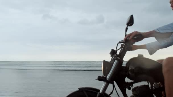 在海滩上骑摩托车的人。复古摩托车在巴厘岛的海滩日落。年轻的时尚男性享受自由和积极的生活方式，有乐趣在自行车之旅 — 图库视频影像
