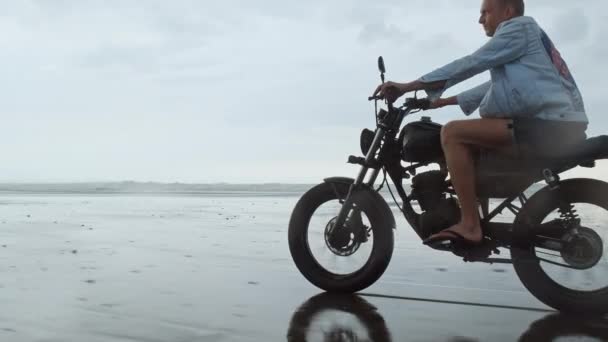 年轻英俊的嬉皮士男子骑着现代定制摩托车赛车手在靠近水的黑色沙滩上。 有海浪的冲浪点. — 图库视频影像