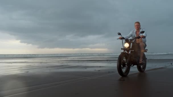 ビーチでオートバイに乗っている男。バリ島のビーチの夕日にヴィンテージバイク。自由とアクティブなライフスタイルを楽しむ若いヒップスターの男性は、バイカーツアーで楽しんでいます — ストック動画