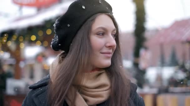 Retrato de una hermosa joven sonriente afuera disfrutando de las nevadas de invierno usando gafas gorra y abrigo mirando a la cámara . — Vídeo de stock
