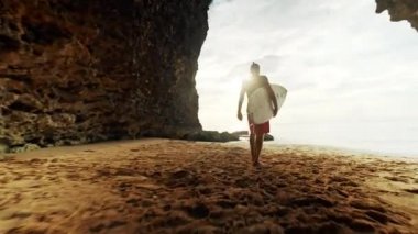 Dalış kıyafeti genç adam sörfçü gün batımında gimbal dolly steadycam okyanusbakarak deniz kıyısında kum üzerinde yürüyüş plajda sörf tahtası alır