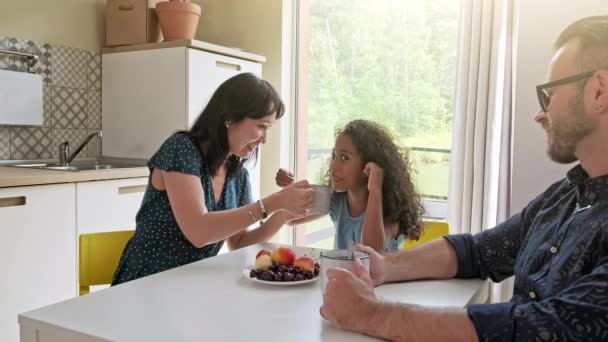 Linda família com criança sentada à mesa bebendo café em uma nova casa em torno de caixas de papelão — Vídeo de Stock