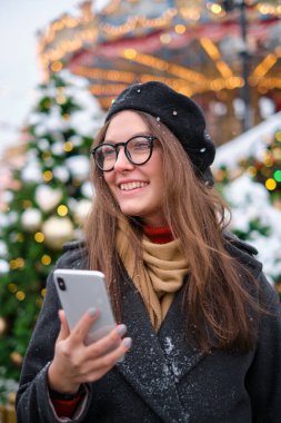 Genç ve çekici bir kız sokakta telefon kullanıyor. Giyinip süslenmiş Noel ağaçlarının arka planında, Noel havasında.,