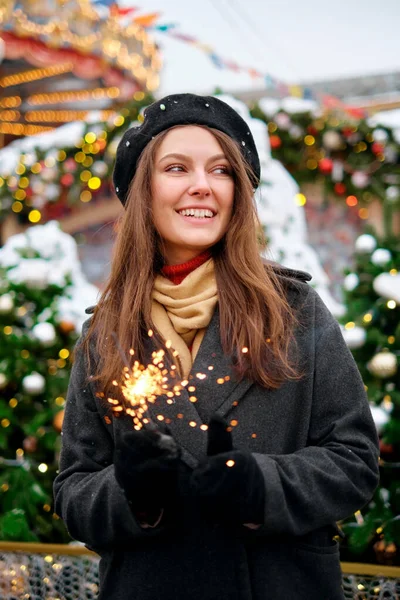 Attractive european lady in beret celebrating new year on the street, holding Bengal light. Outdoor portrait of happy brunette girl posing with sparkler in winter. — Stock Photo, Image