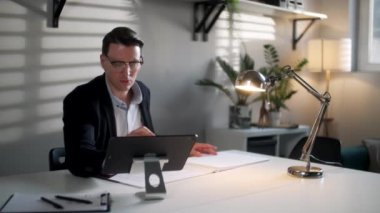 İş adamı mühendisi modern işyerinde planlar ve dizüstü bilgisayarla çalışıyor. Yakışıklı erkek, modern ofis iç işlerinde çalışan, kendine güvenen ve kendine güvenen gözlüklü bir erkek..