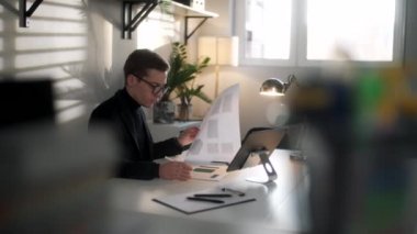 İş adamı mühendisi modern işyerinde planlar ve dizüstü bilgisayarla çalışıyor. Yakışıklı erkek, modern ofis iç işlerinde çalışan, kendine güvenen ve kendine güvenen gözlüklü bir erkek..