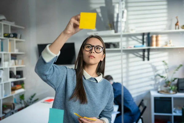 Jonge zakenvrouw aan het brainstormen. Aziatische vrouw het opschrijven van ideeën op plakkerige notities gehecht aan glazen muur. Succesconcept voor bedrijven — Stockfoto