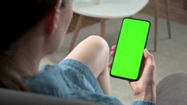 Green Mock-up Smartphone kullanarak Evdeki Genç Kadın 'ın Bir Sandalyede Oturması. Kız, Gadget ekranına dokunmadan İçerik 'i izliyor. Modent Teknoloji ve Bilgi Konsepti.