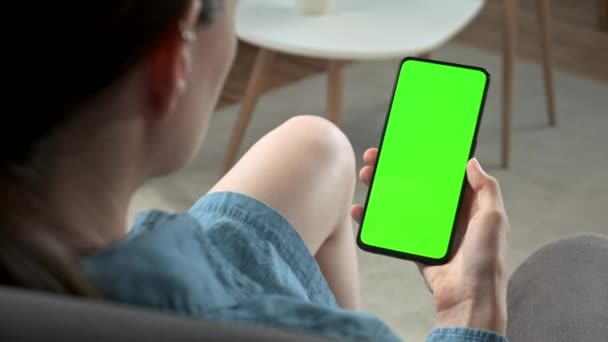 Vista posterior de la mujer joven en casa sentada en una silla con un teléfono inteligente de pantalla falsa verde. Chica está viendo contenido sin tocar la pantalla del gadget. Modent Tecnología y Concepto de Información . — Vídeo de stock