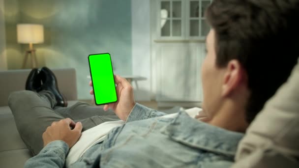 Вид ззаду молодого чоловіка вдома сидить на стільці, використовуючи з зеленим макет екрану смартфона. Хлопчик розміщує контент, не торкаючись екрана гаджетів. Модульна технологія та інформаційна концепція . — стокове відео