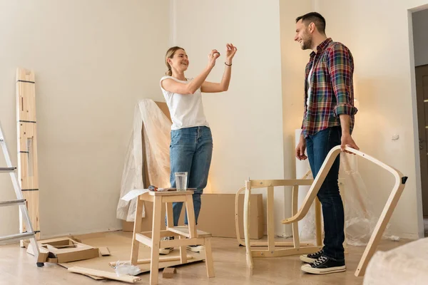 Feliz pareja sonriente recoge muebles como un equipo. Chica ayuda a montar detalles de la silla. Mudándose a Apartamento Nuevo, Joven Familia Montar Muebles — Foto de Stock