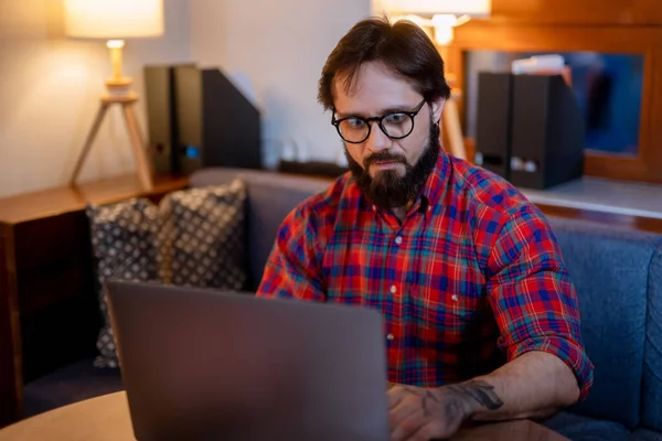 Przystojny mężczyzna w okularach i koszuli, piszący w notatniku, siedzący przy biurku z laptopem w pustym ciemnym biurze.. — Zdjęcie stockowe