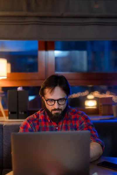 一个戴眼镜，穿着衬衫的英俊男人坐在写字台旁，在空荡荡的黑暗的办公室里拿着笔记本电脑. — 图库照片