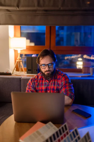一个戴眼镜，穿着衬衫的英俊男人坐在写字台旁，在空荡荡的黑暗的办公室里拿着笔记本电脑. — 图库照片