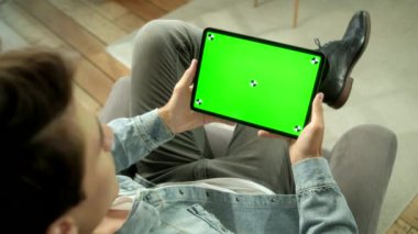 Omuz manzaralı genç adam evde oturuyor yeşil maket tabletini kullanarak bir sandalyede oturuyor. Erkek Dokunmatik Ekran Aygıtı, İnternet Tarayıcı. Yeşil Ekran ve Krom Anahtarı Konsepti.