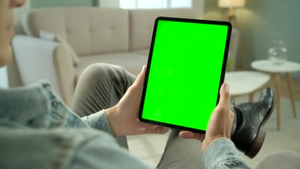 Przez ramię Zobacz młodego człowieka w domu Siedząc na krześle za pomocą zielonej makiety ekranu Tablet. Mężczyzna korzystający z ekranu dotykowego, przeglądający Internet. Koncepcja zielonego ekranu i klucza Chroma. — Wideo stockowe