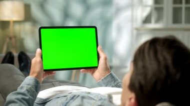 Omuz manzaralı genç adam, yatay modda yeşil maket tablet bilgisayarla kanepeye uzanıyor. Erkek Dokunmatik Ekran Aygıtı Kullanımı, İnternet Taraması,
