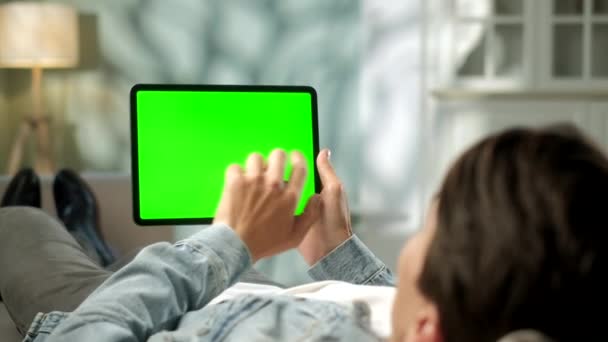 ソファに寝そべっている間、緑のモックアップ画面デジタルタブレットコンピュータで手ジェスチャーを使用して男の肩からの眺め。背景に｜Cozy Living Room. — ストック動画