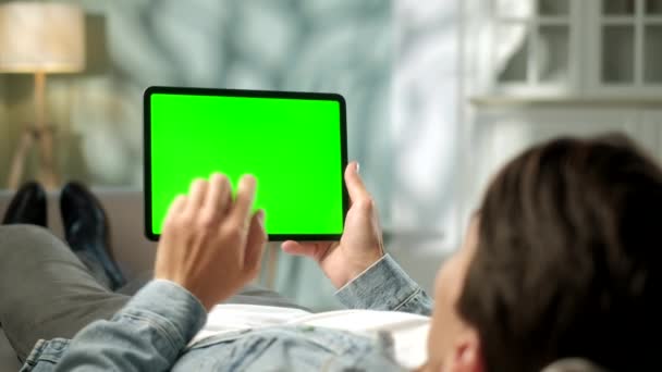 男の肩からの眺めソファの上に横たわる緑のモックアップ画面デジタルタブレットコンピュータ上の手のジェスチャーを保持し、使用しています。男性は物を買うか、インターネットを通して閲覧する. — ストック動画