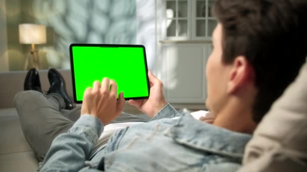 View From the Shoulder of Man Using Hand Gestures on Green Mock-up Screen Digital Tablet Computer in Landscape Mode while Liying on a Sofa. I bakgrunden mysiga vardagsrum. — Stockvideo