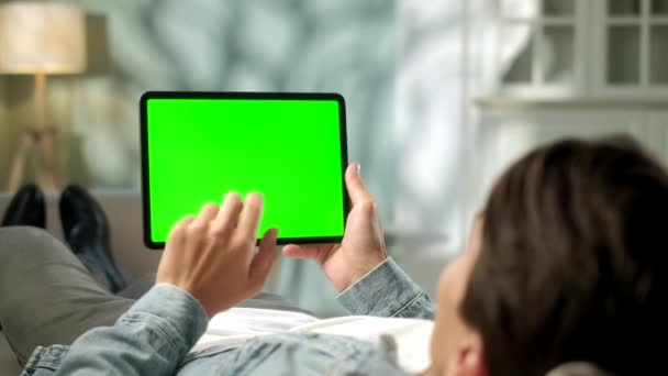 Вид с плеча человека, держащего в руках зеленый макбук-экран и лежащего на софе. Мужчина покупает вещи или просматривает через Интернет . — стоковое видео