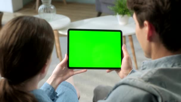 通过肩部视图年轻的夫妇坐在沙发上使用绿色模拟屏幕表格的家庭。男人和女人使用触摸屏设备,浏览互联网.绿色屏幕和色键的概念. — 图库视频影像