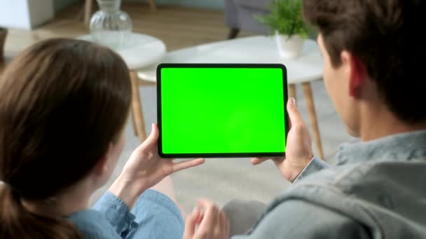 Sobre el hombro Ver joven pareja en casa sentado en un sofá usando con Green Mock-up Screen Tablet. Hombre y mujer utilizando el dispositivo de pantalla táctil, navegar por Internet. Concepto de pantalla verde y clave de croma . — Vídeos de Stock