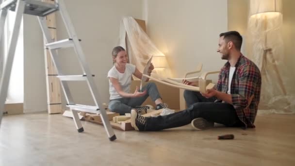 若い大人たちは新しい家に引っ越します。椅子の家具を組み立てるに移動するカップル。新しい家で若い異人種間のカップル。修復、改修、人々の概念は — ストック動画
