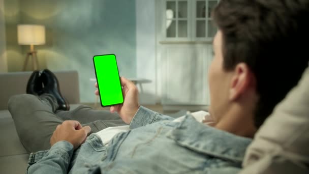 Πίσω όψη του ανθρώπου στο τηλέφωνο με πράσινη οθόνη για αντίγραφο χώρου. Chromakey Mock Up Χωρίς Ανίχνευση Δείκτες. 20άρης βλέποντας ειδήσεις βίντεο στον καναπέ από κοντά. Πατήστε για να κάνετε κλικ στο κέντρο της οθόνης — Αρχείο Βίντεο