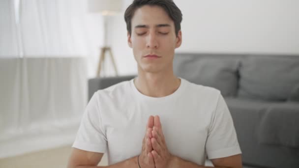 El joven relajado en ropa deportiva está meditando en posición de loto sentado en una esterilla de yoga con los ojos cerrados en casa disfrutando de la meditación con las manos en Namaste . — Vídeo de stock
