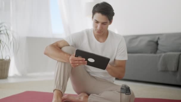 Sonriente joven navegando por Internet en la tableta mientras está sentado en la esterilla de ejercicio durante la relajación después del entrenamiento en casa . — Vídeo de stock
