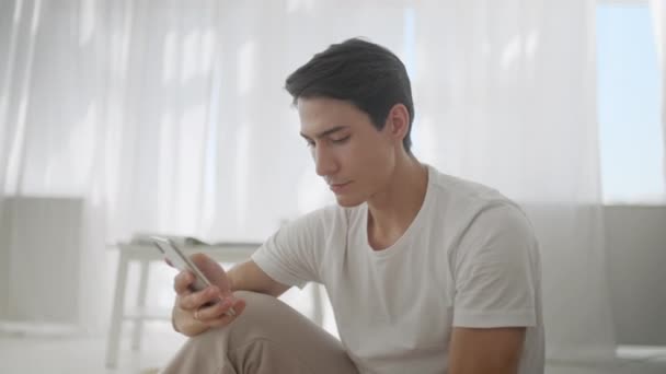 Glimlachende jonge man surfen op internet via de telefoon tijdens het zitten op Exercise Mat tijdens ontspannen na het sporten thuis. — Stockvideo