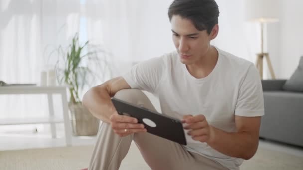 Sorrindo Jovem Surfando na Internet em Tablet Enquanto Sentado no Tapete de Exercício Durante o Relaxamento Depois do Treino em casa . — Vídeo de Stock