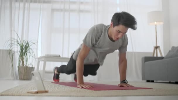 Hombre joven guapo con una camiseta gris y pantalones cortos negros hace deporte en el hogar en línea. Hombre guapo haciendo ejercicio y haciendo flexiones en la sala de estar en casa . — Vídeo de stock