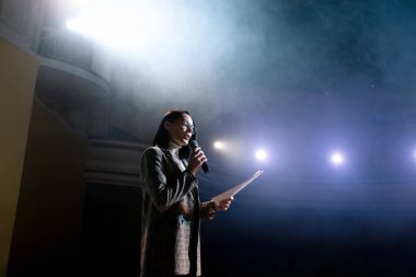 Kadın finans koçu, forumda seyircilerle sahnede konuşurken duygusal jestler yapıyor. Birçok isimsiz insan ve öğrenci büyük bir salonda oturup atölye çalışmalarını izliyor.