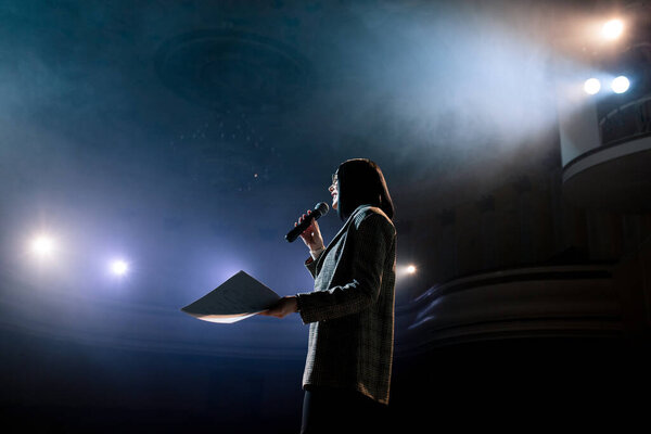 Женщина-финансовый тренер эмоционального жеста разговаривает со зрителями на форуме со сцены. Слишком много анонимных работников и студентов сидят в большом зале и смотрят семинар фон
