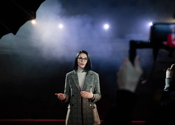 Show woman. Female entertainer, presenter or actor on stage. Arms to sides, smoke on background of spotlight. Front view portrait of a female public speaker speaking at the microphone, pointing