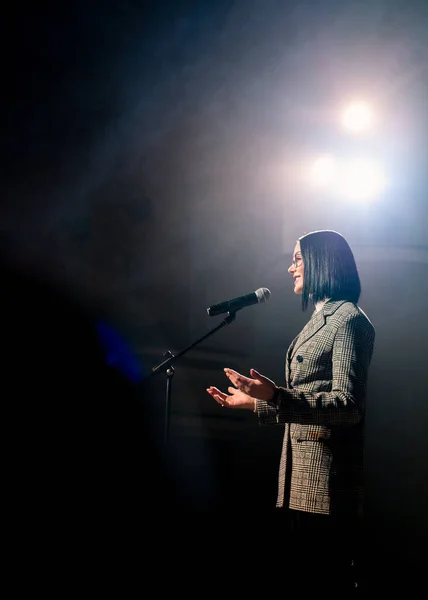 Show woman. Female entertainer, presenter or actor on stage. Arms to sides, smoke on background of spotlight. Front view portrait of a female public speaker speaking at the microphone, pointing