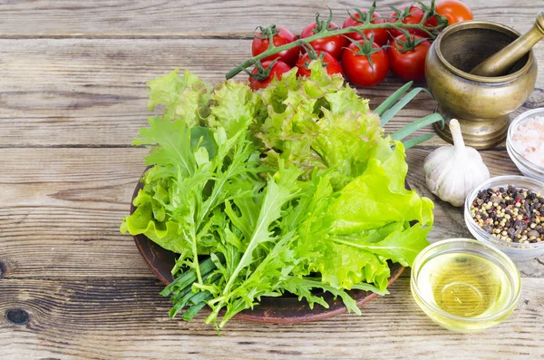 Green salad ingredients organic lettuce, cherry tomatoes, spices and olive oil on wooden background.