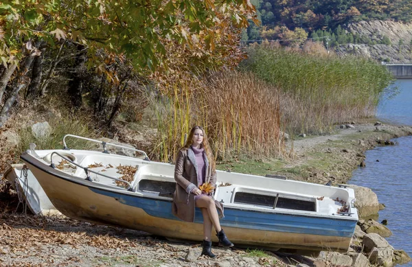 Young beautiful woman with leaves in hands sits on a boat by the lake in the mountains on a autumn, sunny day close-up