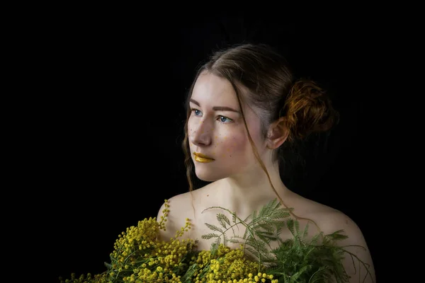 Portrait of a young, beautiful woman with a bouquet of yellow, spring mimosa on a black background close-up.