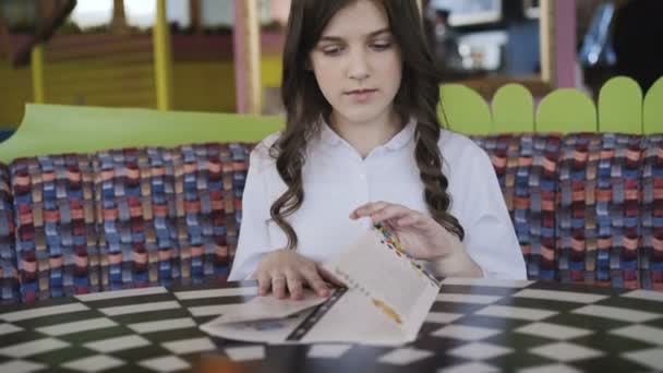 Menina bonita, sorridente e jovem lendo o menu com maravilha no café. 4K — Vídeo de Stock