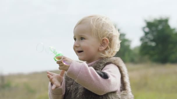 Портрет счастливого, улыбающегося малыша надувает пузырьки на природе. — стоковое видео
