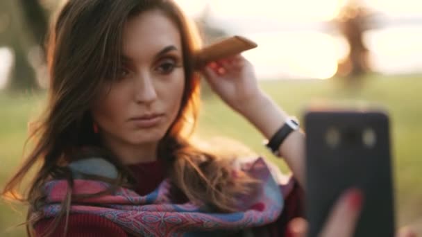 Chica feliz peinando el pelo en el teléfono inteligente en un parque de otoño — Vídeos de Stock