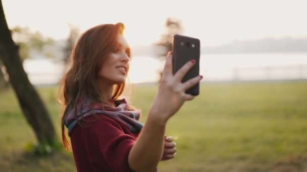 Chica positiva sonriendo y posando en el teléfono inteligente para selfies en otoño soleado parque — Vídeos de Stock