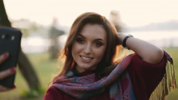Chica despreocupada sonriendo y posando en el teléfono inteligente para selfies en otoño soleado parque — Vídeos de Stock
