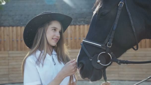 Schattig meisje in de hoed komt en streelt zwarte merries hoofd op zonnige achtergrond — Stockvideo