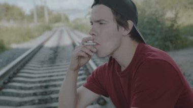 Genç düşünceli çocuk bir demiryolu üzerinde oturur ve sigara içiyor. 4k