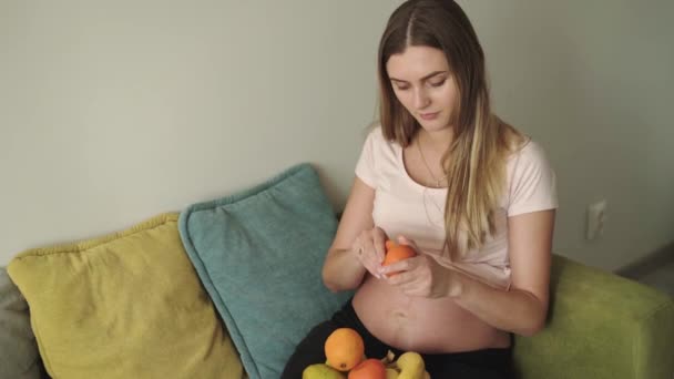Hambrienta mujer embarazada pelando mandarinas corteza en el sofá en casa — Vídeo de stock